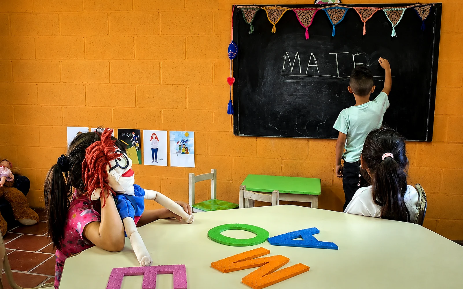 Un chico escribe MATEO en el pizarrón mientras sus compañeros trabajan con letras móviles sobre la mesa